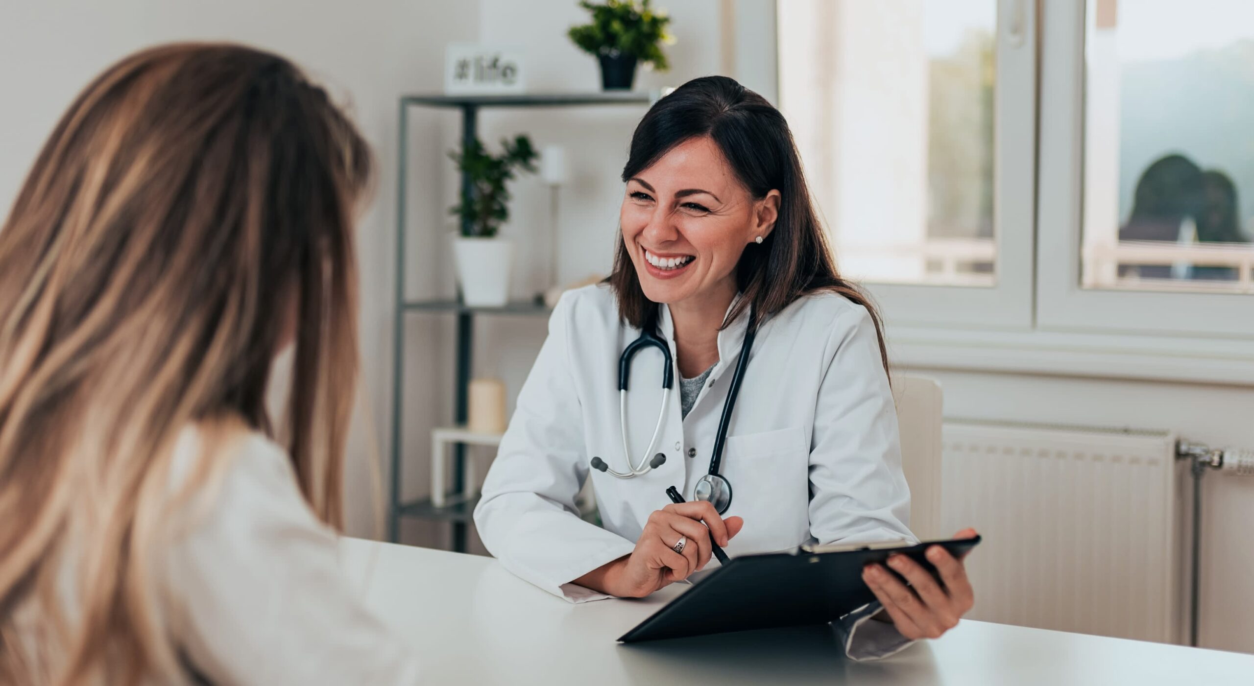 IXXDNA Smiling clinician in a white coat with a stethoscope talks with a woman across a desk, taking notes on a clipboard in a bright clinic office.
